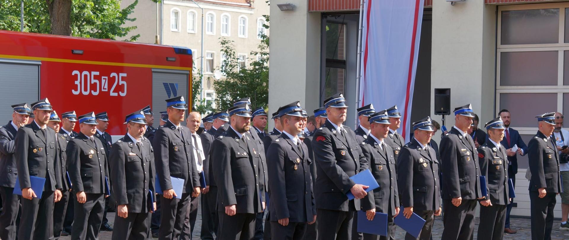 Na zdjęciu widoczni są strażacy druhowie wraz z niebieski teczkami - grantami. W tle widać budynek Komendy Wojewódzkiej PSP w Szczecinie oraz samochód ratowniczo-gaśniczy
