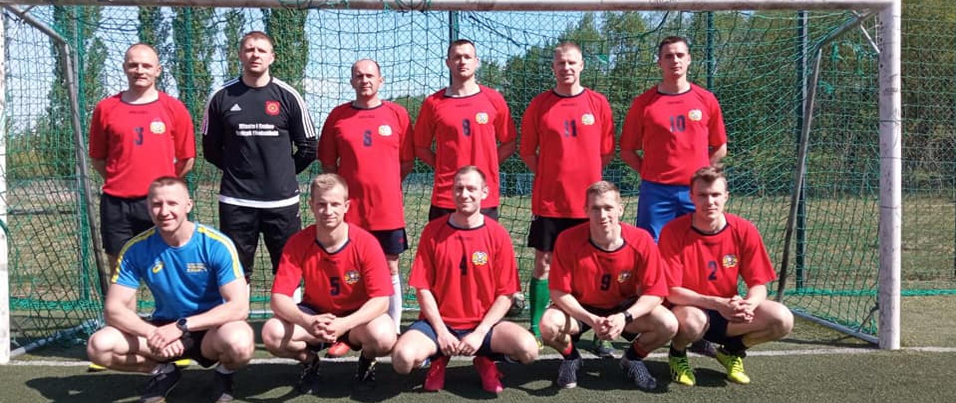 Zdjęcie przedstawia 11-osobową męska drużynę piłkarską pozującą do zdjęcia w czerwonych strojach sportowych na tle bramki piłkarskiej.