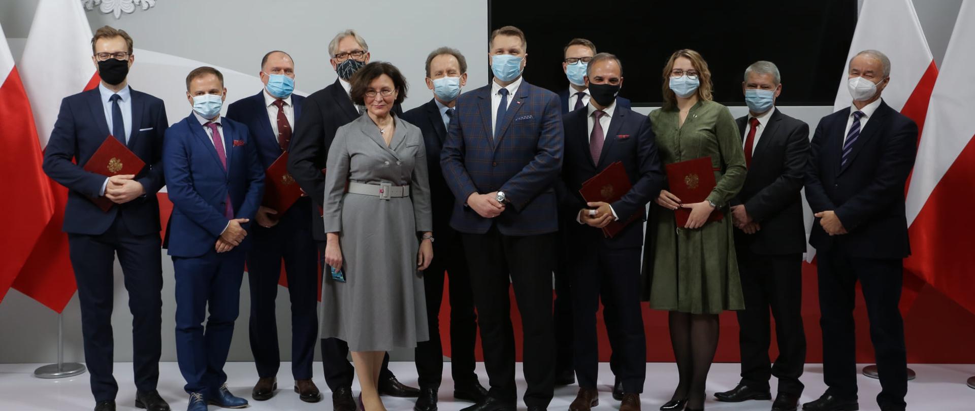 Grupa osób trzymających w ręku czerwone teczki. Wśród nich pośrodku znajduje się minister Przemysław Czarnek oraz za jego plecami wiceminister Włodzimierz Bernacki. Większość z osób ma na twarzy maseczki. W tle monitor telewizyjny i biało-czerwone flagi.