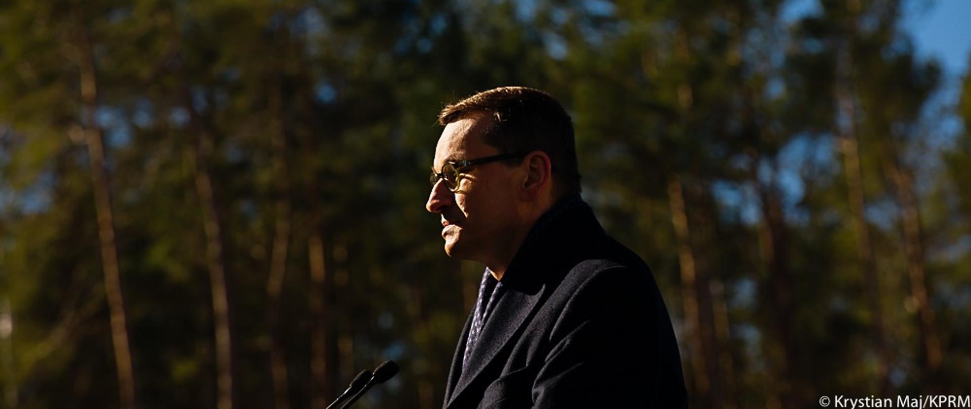 Premier Mateusz Morawiecki przy mikrofonie. W tle drzewa.