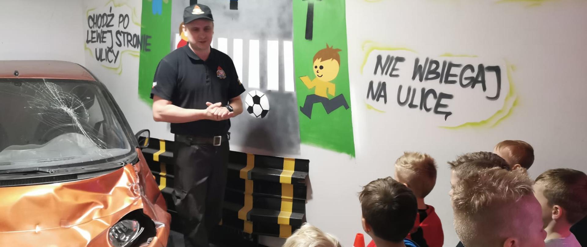 Zdjęcie przedstawia grupę dzieci słuchających strażaka, który opowiada o zagrożeniach na stanowisku imitującym wypadek drogowy z rozbitym autem osobowym.