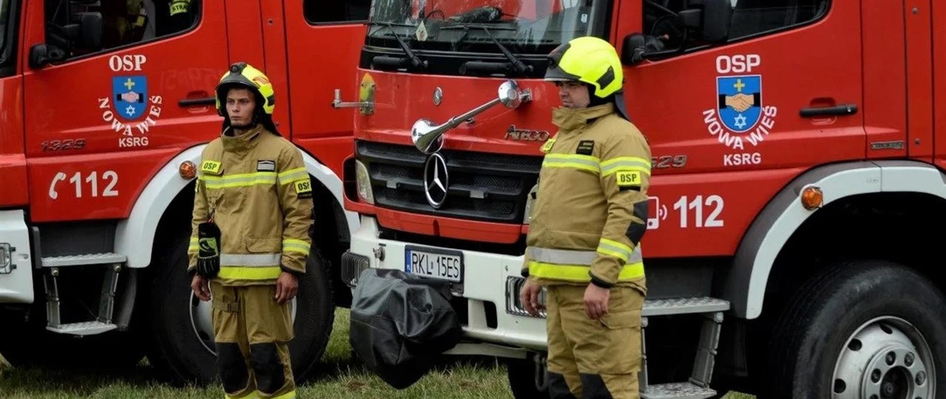 Na trawie stoją częściowo widoczne dwa samochody strażackie widziane z przodu i boku. Przy jednym z nich po prawej i lewej stronie stoją strażacy w umundurowaniu bojowym w kolorze piaskowym.