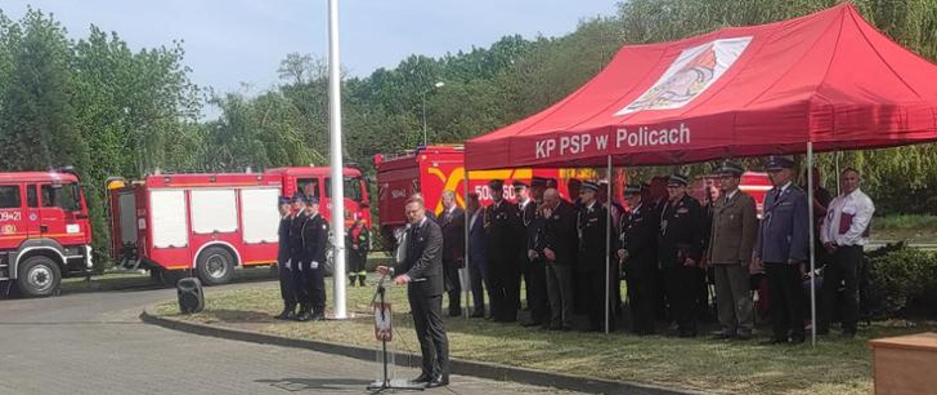 Na zdjęciu trzech strażaków w galowych mundurach z żółtymi sznurami i rogatywkach na głowie stojących przy białym maszcie. Obok widoczna przejrzysta mównica z godłem narodowym i przemawiający Wojewoda zachodniopomorski Pan Zbigniew Bogucki. Za Nim czerwone zadaszenie otwartego namiotu z napisem KP PSP w Policach i logiem Państwowej Straży Pożarnej, pod którym widać stojące postaci, część z nich w mundurach. W głębi widoczne prawe boki pojazdów pożarniczych, zieleń drzew i krzewów, błękitne niebo.
