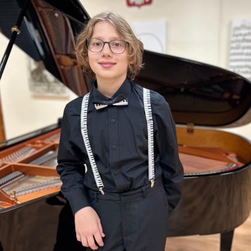 Zdjęcie przedstawia chłopca w okularach ubranego na czarno i opartego o czarny fortepian. W tle rozmazane grafiki o tematyce muzycznej. 