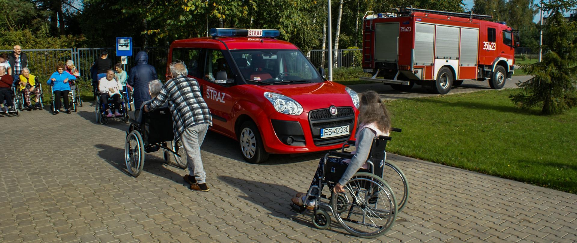 Na zdjęciu widać plac przed budynkiem domu pomocy społecznej na którym gromadzą się osoby ewakuowane. Są to mieszkańcy domu pomocy społecznej, którzy gromadzą się na parkingu w wyznaczonym miejscu. Częśc z osób porusza się na wózkach inwalidzkich. W tle widać pojazdy straży pożarnej. Pogoda słoneczna. W ewakuacji osób na wózkach pomagają pracownicy domu pomocy społecznej.