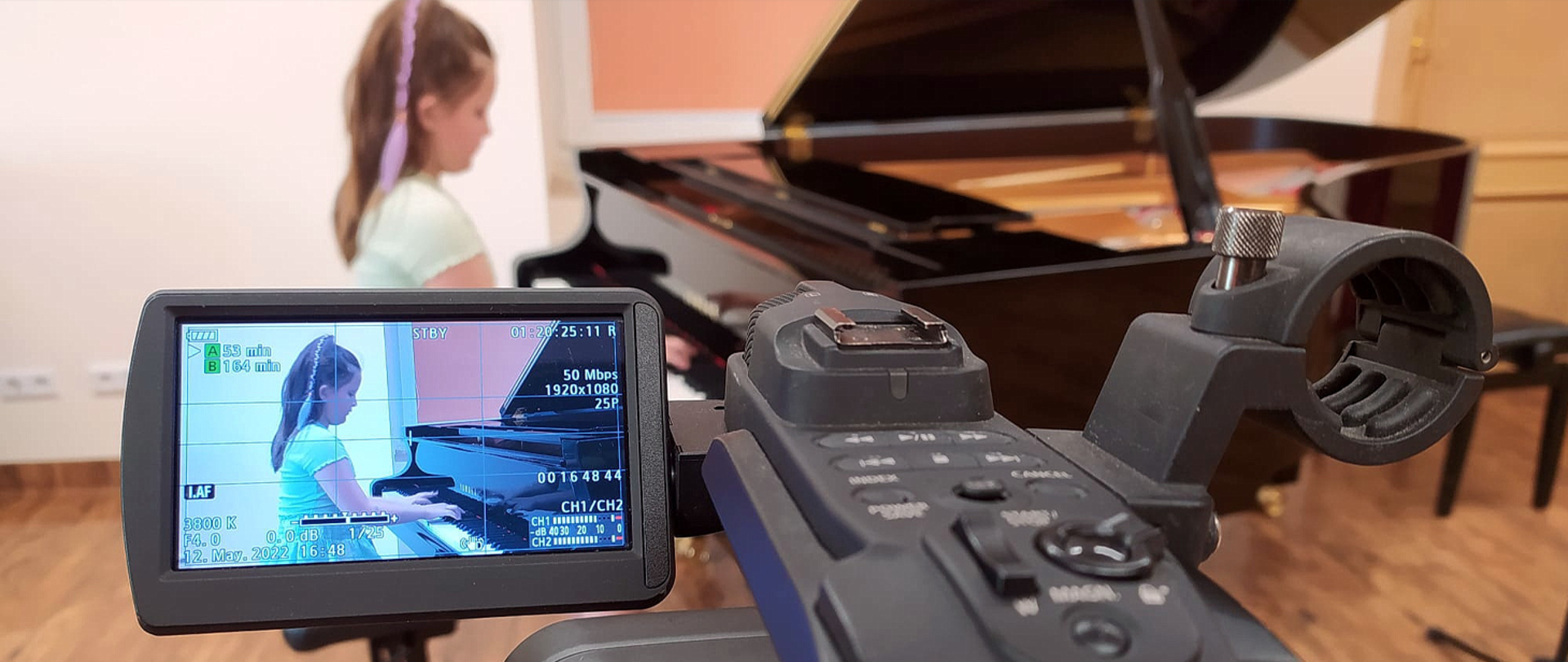 Na zdjęciu w tle rozmyty widok dziewczynki grającej na fortepianie, na pierwszym planie ostry obraz ekranu LCD kamery telewizyjnej i fragmentu samej kamery nagrywającej widoczną w tle dziewczynkę.