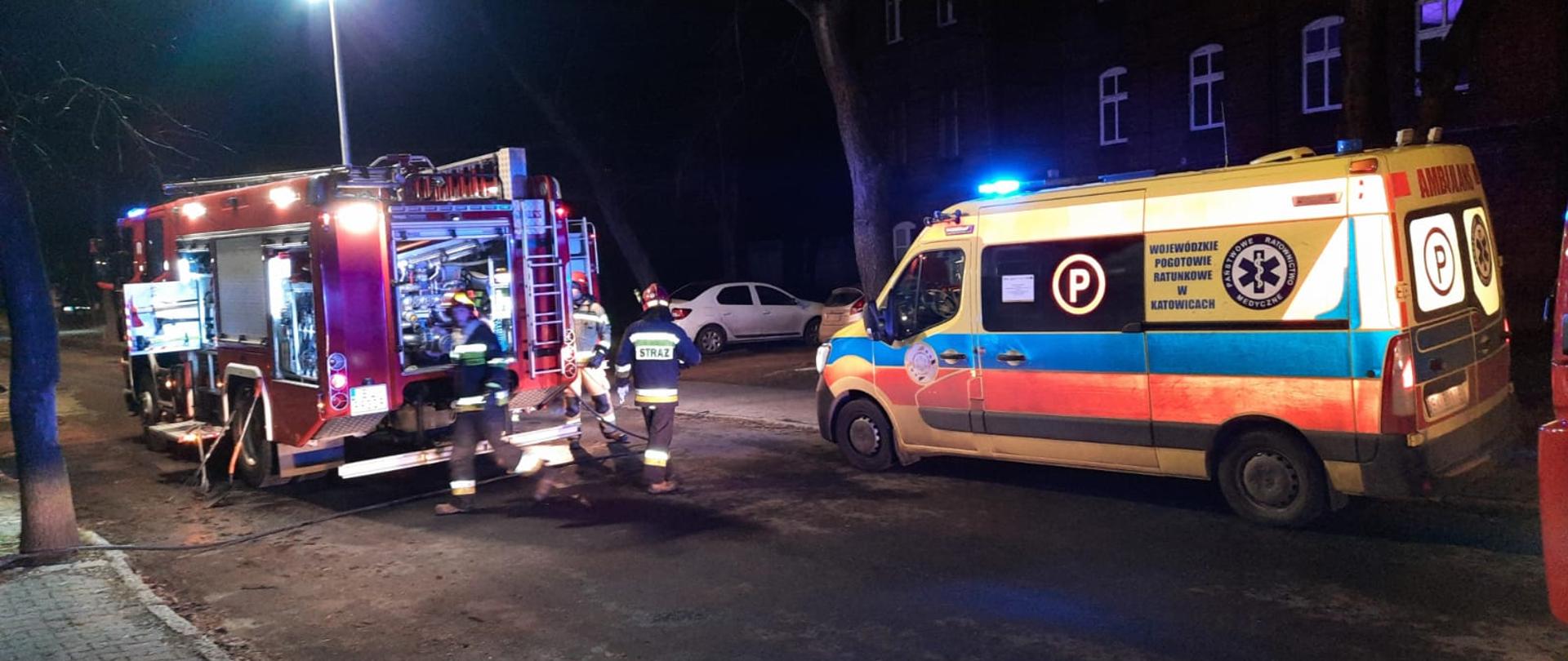 Zdjęcie przedstawia budynek, w którym wybuchł pożar oraz strażaków, pracujących na miejscu zdarzenia. Na zdjęciu również widnieje zespół ratownictwa medycznego.