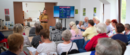 Zdjęcie przedstawia seniorów zebranych na sali, siedzą przed stołami, przed nimi stoi strażak i przedstawia informacje dotyczące czujek