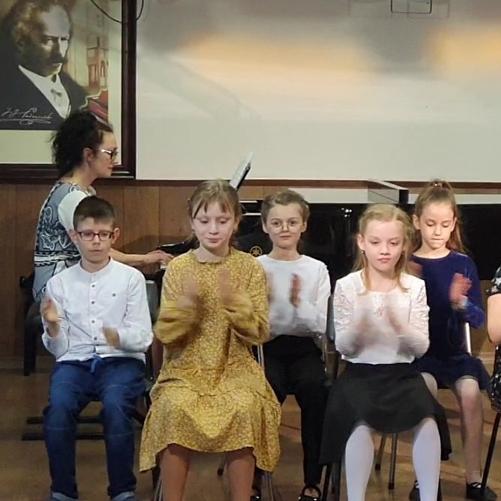 W centrum klaszczący uczniowie ubrani na galowo siedzą na krzesełkach ustawionych na scenie, za nimi z lewej strony przy fortepianie siedzi kobieta w czarnych włosach w okularach. Ściany koloru białego. Na tylnej ścianie wisi obraz z wizerunkiem Paderewskiego.