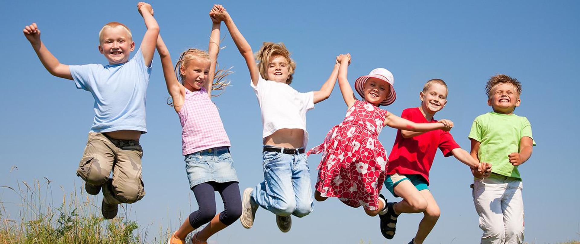 Na zdjęciu grupa dzieci. Skacząc trzymają się za ręce. 
