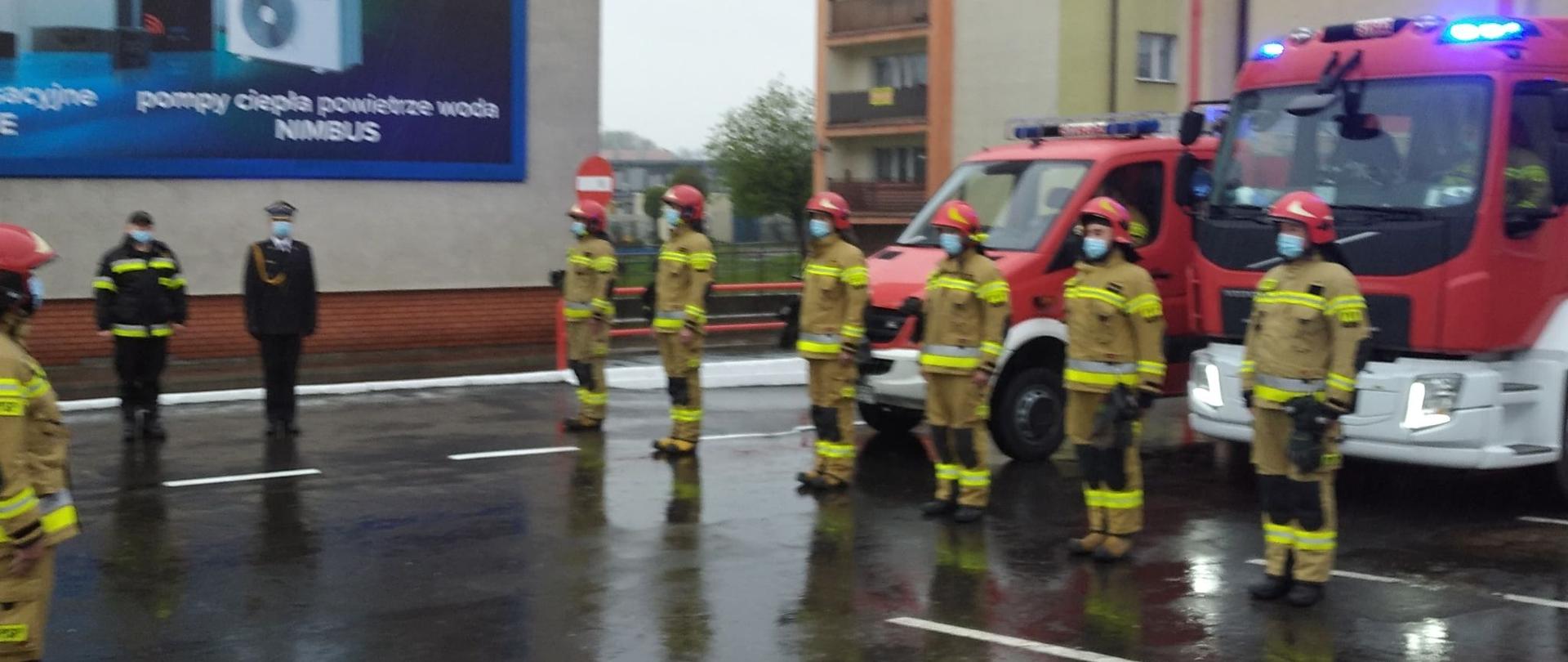 Zdjęcie przedstawia funkcjonariuszy Państwowej Straży Pożarnej, w trakcie uroczystej zmiany służby, w tle dwa pojazdy ratownicze.