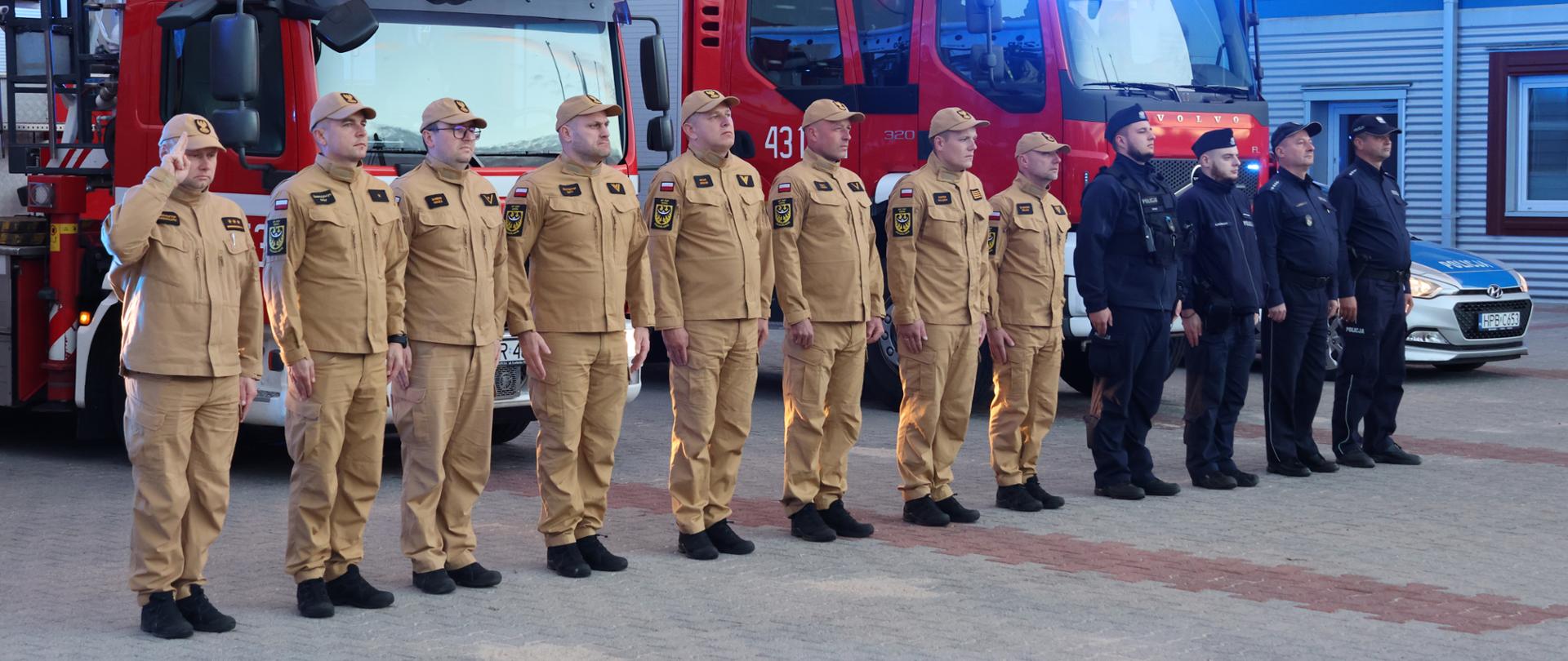Na zdjęciu przedstawiono funkcjonariuszy Państwowej Straży Pożarnej w piaskowych ubraniach służbowych oraz funkcjonariuszy Policji w ciemnych mundurach, stojących w szeregu przed budynkiem straży pożarnej. W tle widoczne są czerwone samochody pożarnicze, w tym pojazd z drabiną mechaniczną, oraz oznakowany radiowóz policyjny. Na dachu pojazdów strażackich włączone są niebieskie sygnały świetlne.