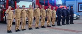 Na zdjęciu przedstawiono funkcjonariuszy Państwowej Straży Pożarnej w piaskowych ubraniach służbowych oraz funkcjonariuszy Policji w ciemnych mundurach, stojących w szeregu przed budynkiem straży pożarnej. W tle widoczne są czerwone samochody pożarnicze, w tym pojazd z drabiną mechaniczną, oraz oznakowany radiowóz policyjny. Na dachu pojazdów strażackich włączone są niebieskie sygnały świetlne.