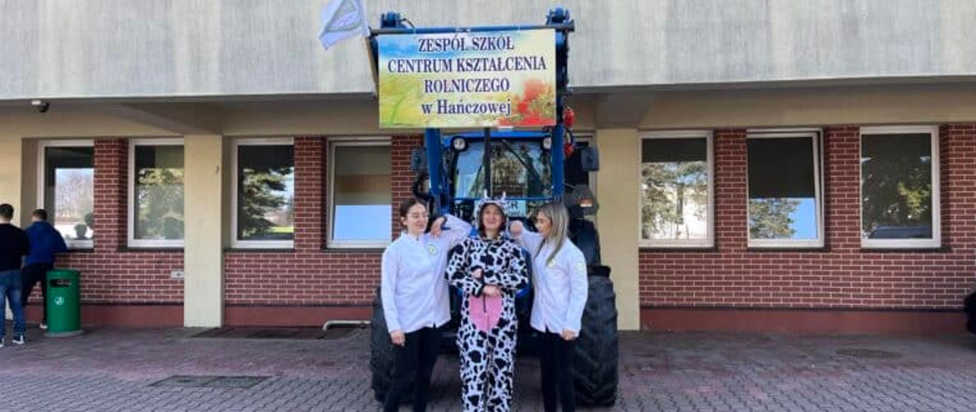 Trzy uczennice przed niebieskim ciągnikiem z banerem ‘Zespół Szkół CKR w Hańczowej’ na dziedzińcu szkoły.