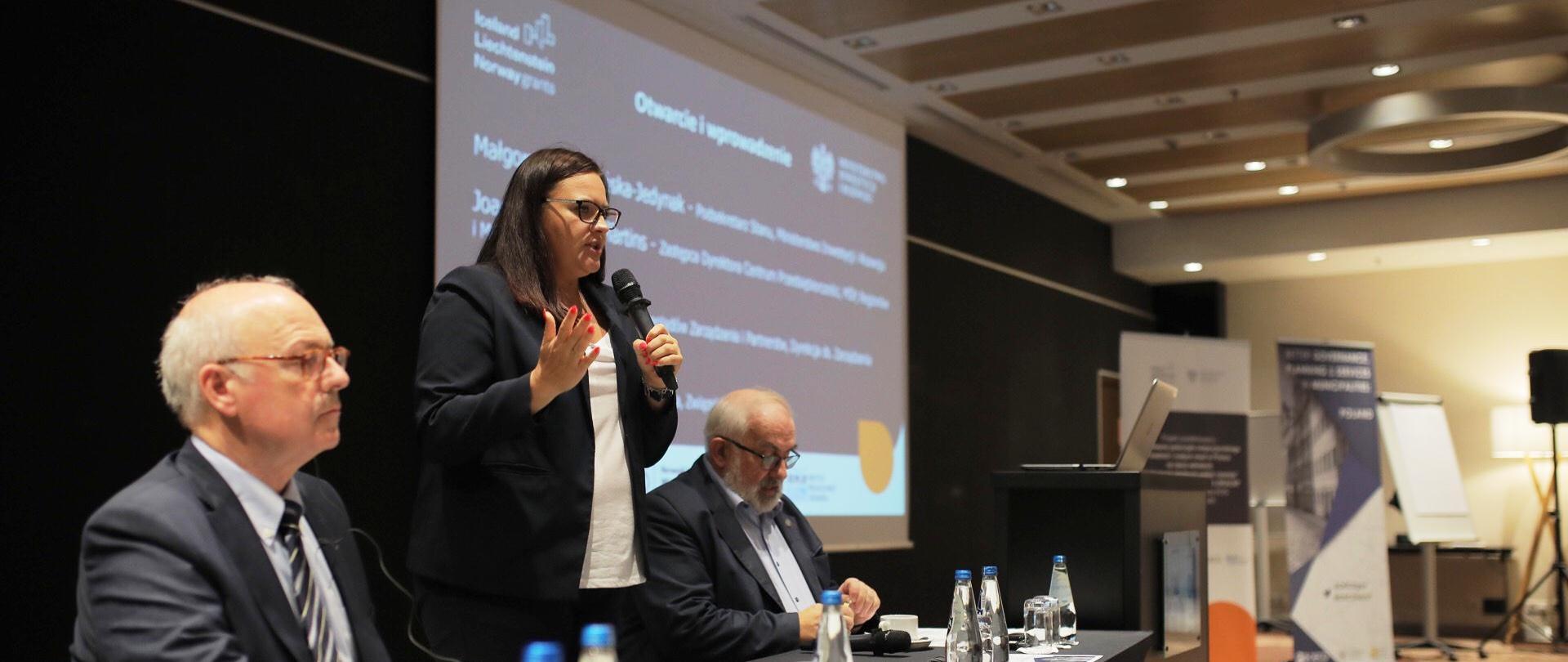 Wiceminister Jarosińska-Jedynak podczas otwarcia konferencji przy stole prezydialnym, obok niej dwie osoby, za nimi ekran z logotypami ministerstwa i funduszy norweskich, na dalszym planie roll-upy.