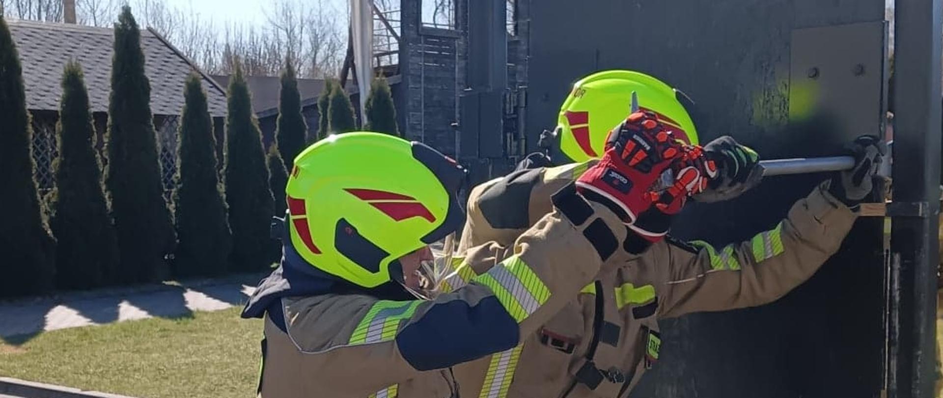 Na zdjęciu widzimy dwóch strażaków podczas próby siłowego otwarcia drzwi z użyciem sprzętu burzącego.