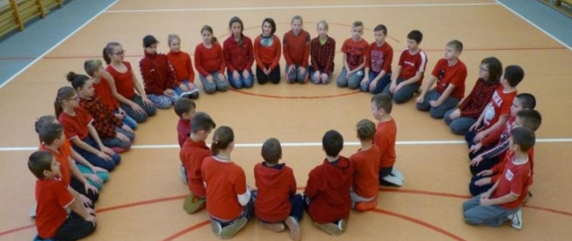 Dzieci w czerwonych koszulkach siedzące w kształcie logo polskiej pomocy w sali gimnastycznej
