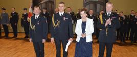 Dzień Strażaka w Rawiczu. Sala widowiskowa w domu kultury. Na tle pododdziału w szeregu stoją: komendant powiatowy PSP, strażak wyróżniony nagrodą komendanta wojewódzkiego, pani minister oraz komendant wojewódzki PSP.