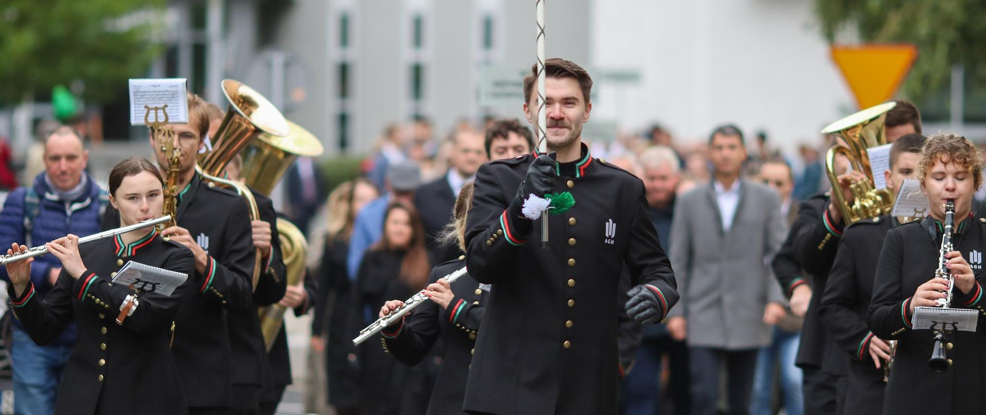 Duża grupa ludzi idzie ulicą, na czele człowiek z elegancką laską i orkiestra w galowych czarnych strojach.