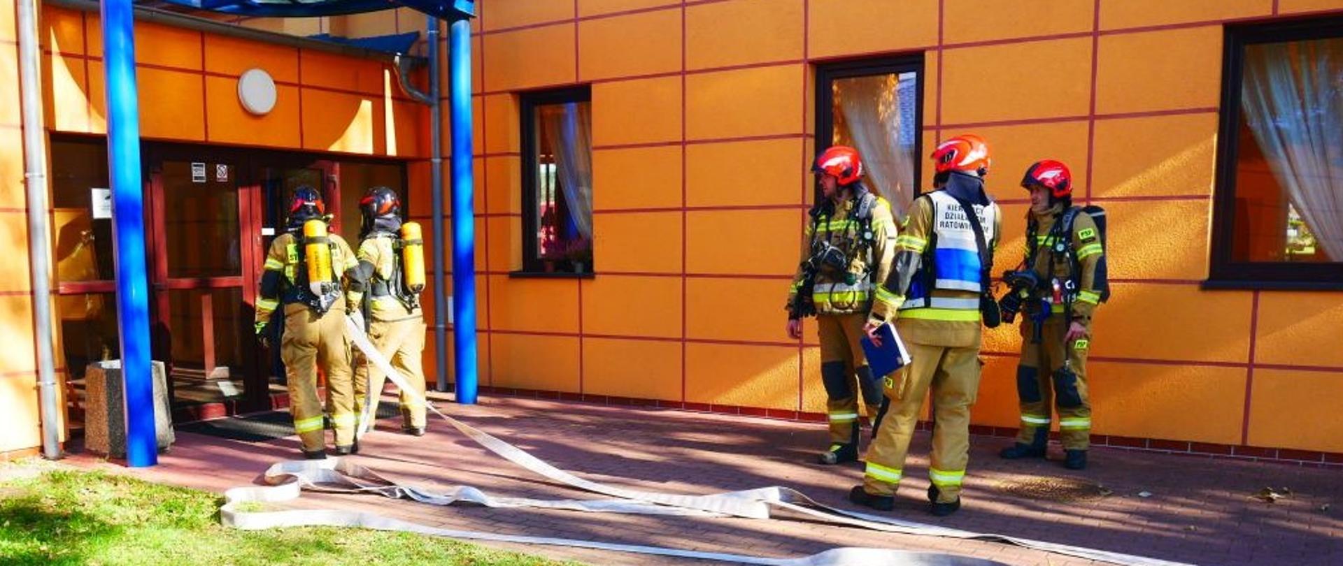 wchodzący do budynku ratownicy w aparatach ochrony dróg oddechowych, obok kierujący akcją ratowniczą i rota zabezpieczająca 