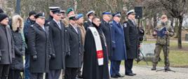 Przedstawiciele służb mundurowych oraz zaproszeni goście stoją obok siebie niedaleko stoi żołnierz przy mikrofonie i czyta.