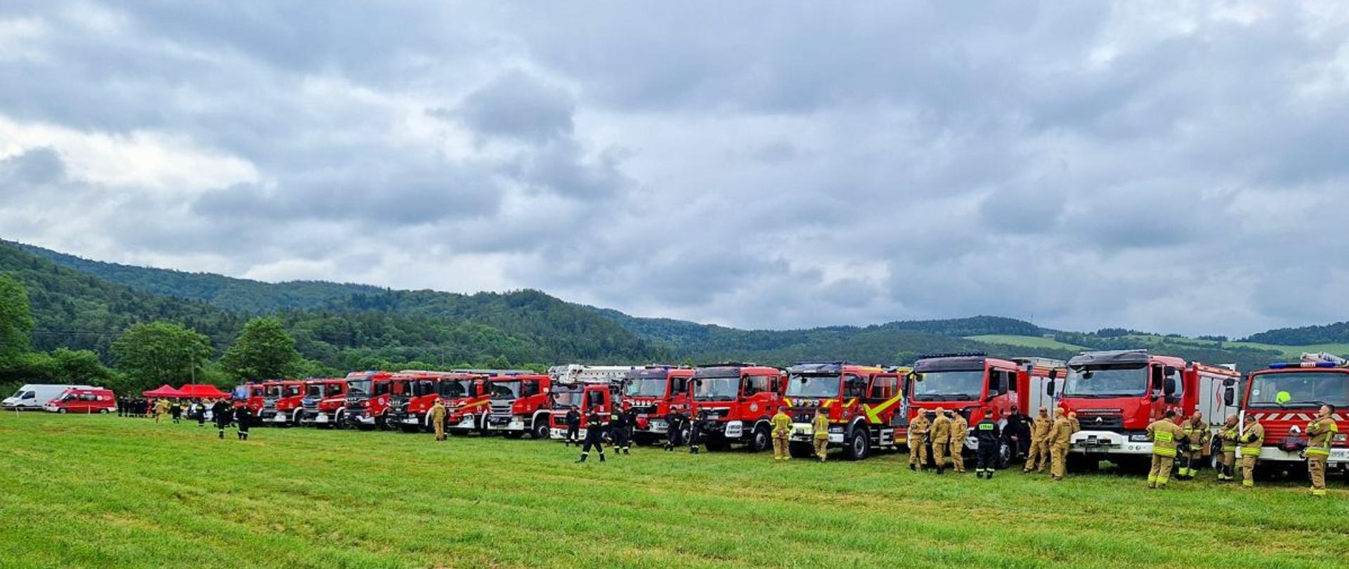 Zdjęcie przedstawia ustawione w rzędzie samochody gaśnicze biorące udział w ćwiczeniach pod kryptonimem "SANOK 2025". Na zdjęciu widać również strażaków którzy stoją przy samochodach. 