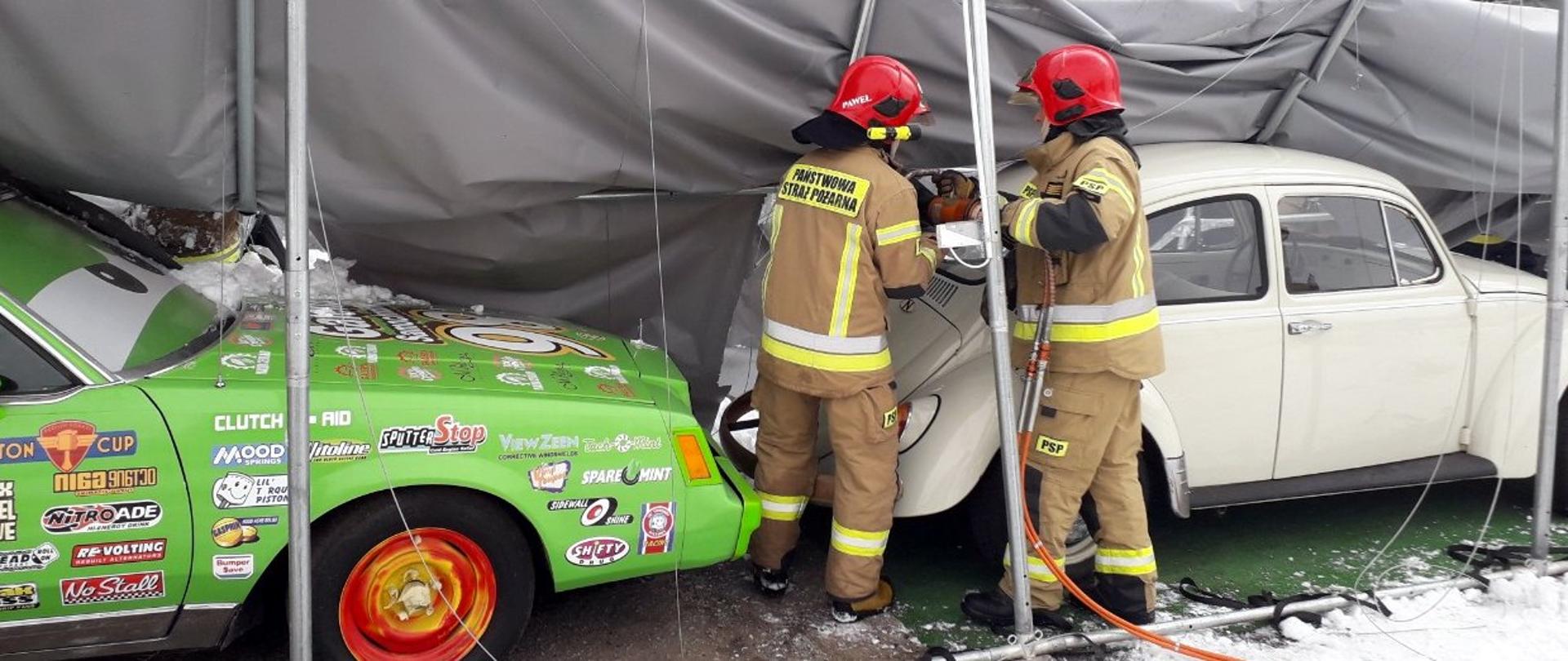 Na zdjęciu dwóch strażaków rozbierających aluminiową konstrukcję uszkodzonego namiotu. Namiot pokryty brezentem. Pod częściowo rozebranym namiotem znajdują się dwa osobowe samochody. Po lewej stronie zdjęcia ujęto przód zielonego samochodu. Karoseria oklejona jest naklejkami. Po prawej stronie widać volkswagena garbusa. Strażacy ujęci są w centrum zdjęcia.