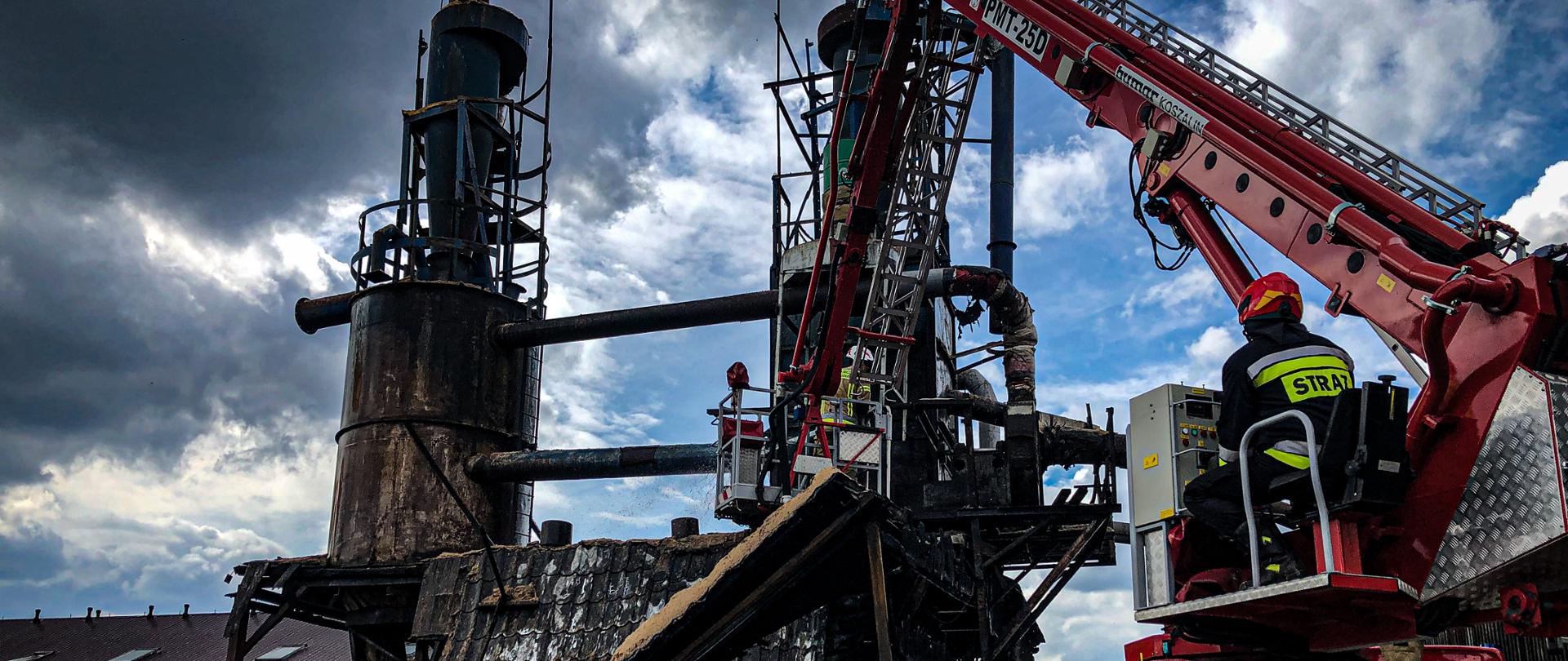 W prawej części fotografii znajduje się rozłożony podnośnik strażacki z koszem w którym znajdują się strażacy. Po lewej stronie fotografii znajdują się rozsypane trociny, a za nimi znajduje się zawalona wiata drewniana z dwoma wystającymi silosami. W oddali znajdują się budynki oraz obok budynków składowane są palety. Zdjęcie wykonano w dzień przy zachmurzeniu.