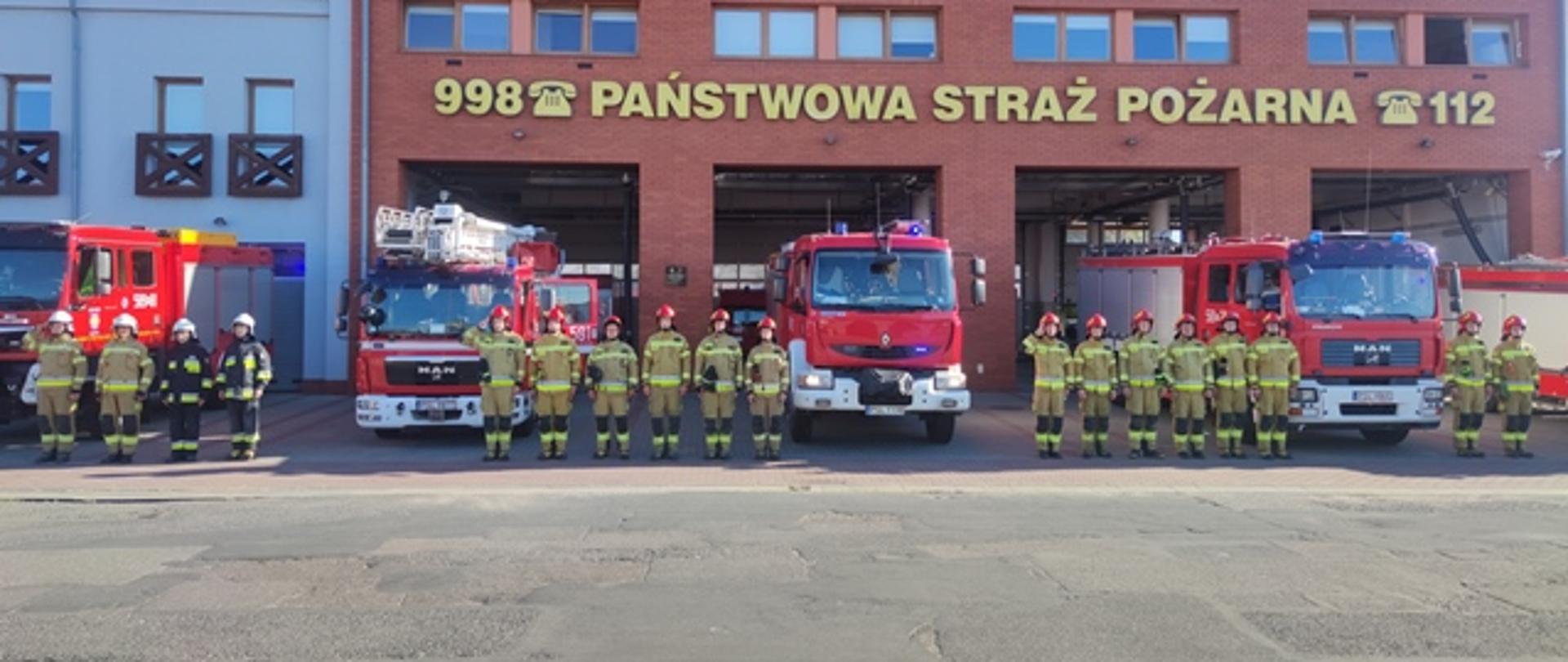 Na tle budynku Komendy samochody pożarnicze PSP i OSP z włączonymi sygnałami przed samochodami strażacy w mundurach bojowych, w szeregu poszczególni dowódcy salutują. 