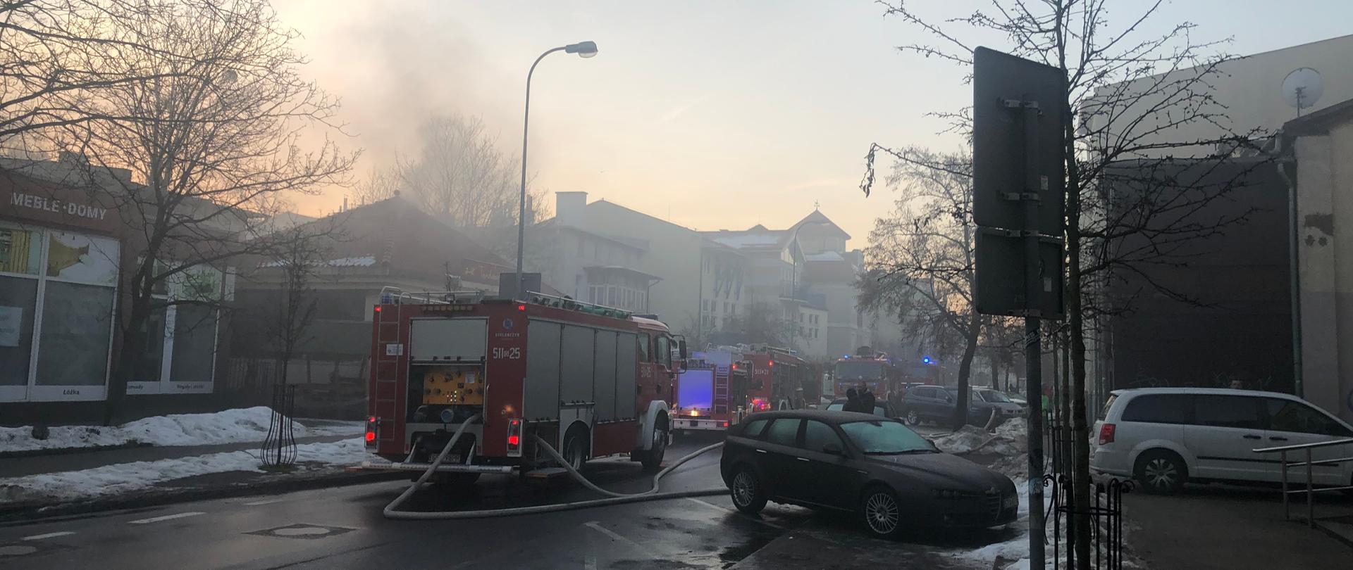 Budynek parterowy z którego wydobywa sie dym. Na ulicy przed budynkiem stoi 5 samochodów strażackich gaśniczych. 