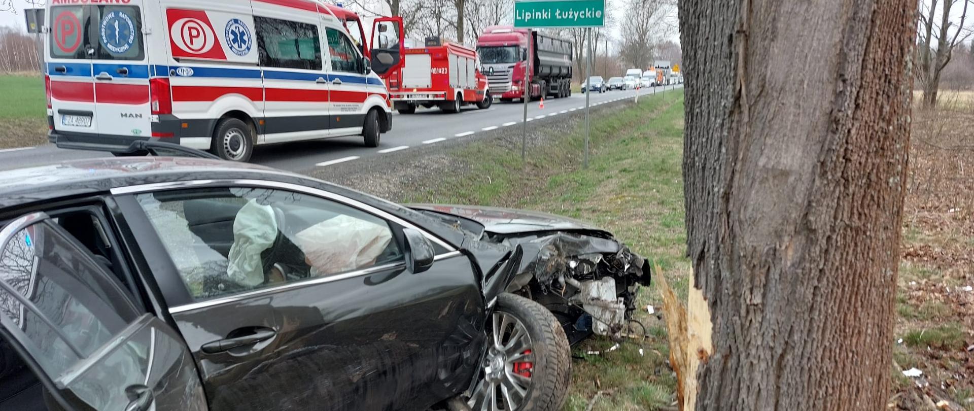 Na zdjęciu widać uszkodzony samochód osobowy marki Mazda po zderzeniu z przydrożnym drzewem znajdującego się na poboczu drogi krajowej numer dwadzieścia siedem. wyniku zderzenia uszkodzony jest przód i bok od strony pasażera samochodu. Na drodze przy samochodzie znajdują się karetka pogotowia i samochód ratownictwa technicznego straży pożarnej. Droga zablokowana. 