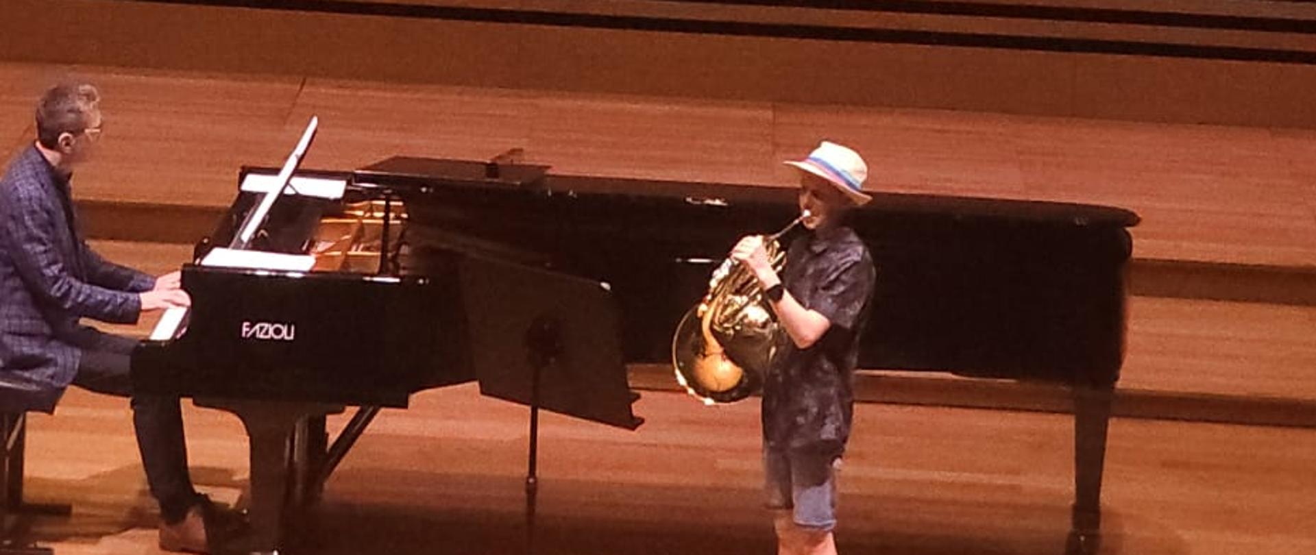 Chłopiec gra na waltorni, mężczyzna akompaniuje na fortepianie.