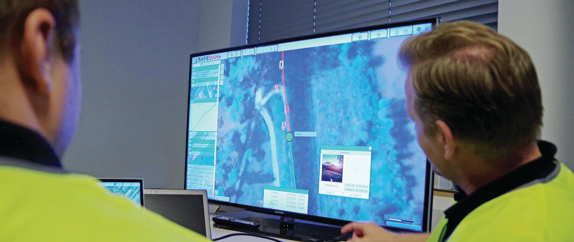 Dwóch mężczyzn w żółtych kamizelkach odblaskowych śledzi na monitorze komputera dane z systemu SAFEDAM
