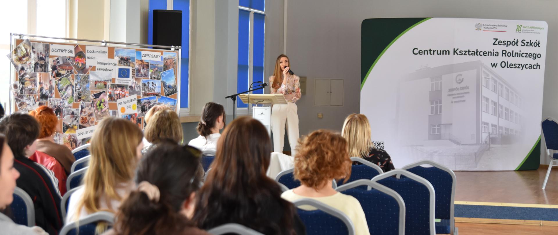 Pani Adrianna Wysocka, psycholog i psychoterapeuta, przedstawia prezentację o zaburzeniach odżywiania, w tle uczniowie i nauczyciele w auli. 