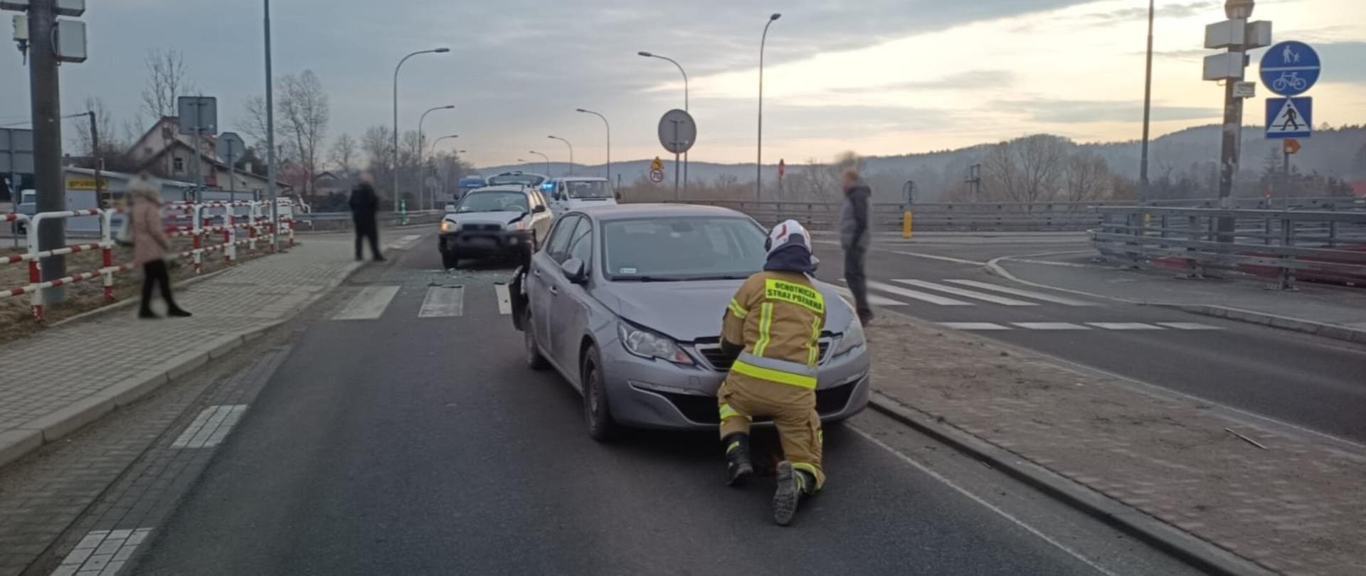 Strażak przed samochodem uczestniczącym w zdarzeniu otwiera klapę silnika w tele drugi samochód uczestniczący w zdarzeniu.