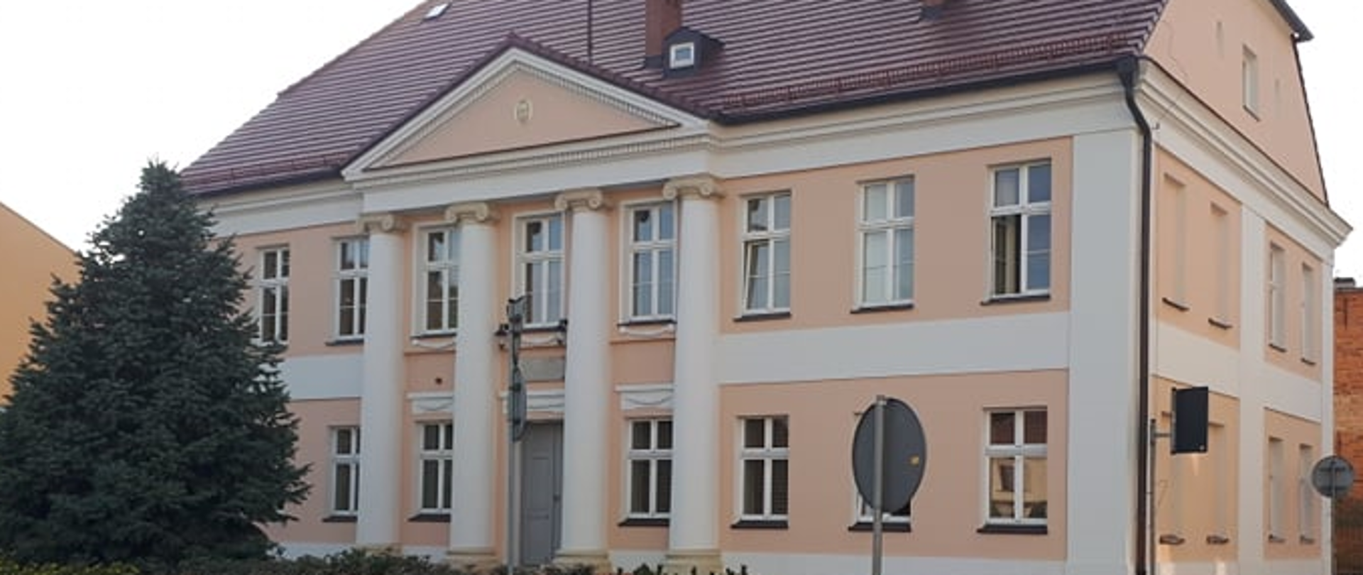 Widoczny jest front oraz ściana boczna budynku. Na froncie znajdują się cztery kolumny rozdzielone oknami, na parterze między drugą a trzecią kolumną są drzwi główne do budynku. Przed budynkiem z lewej strony znajduje się wysokie drzewo iglaste a w dole na środku klomb zieleni.