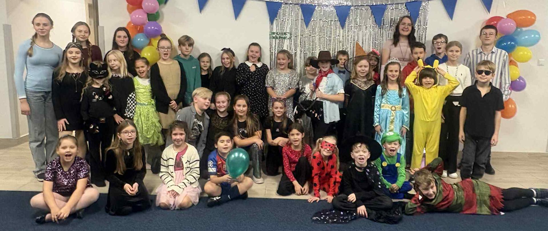 Grupa dzieci bawi się i tańczy w sali podczas szkolnej zabawy kostiumowej. Widać różnorodne stroje, m.in. błękitną suknię księżniczki, żółty kostium, strój czarownicy i przebranie biedronki. Dzieci podskakują i machają rękami w rytm muzyki. Sala jest oświetlona delikatnymi kolorowymi światłami, a w tle widać okna i wiszące dekoracje.