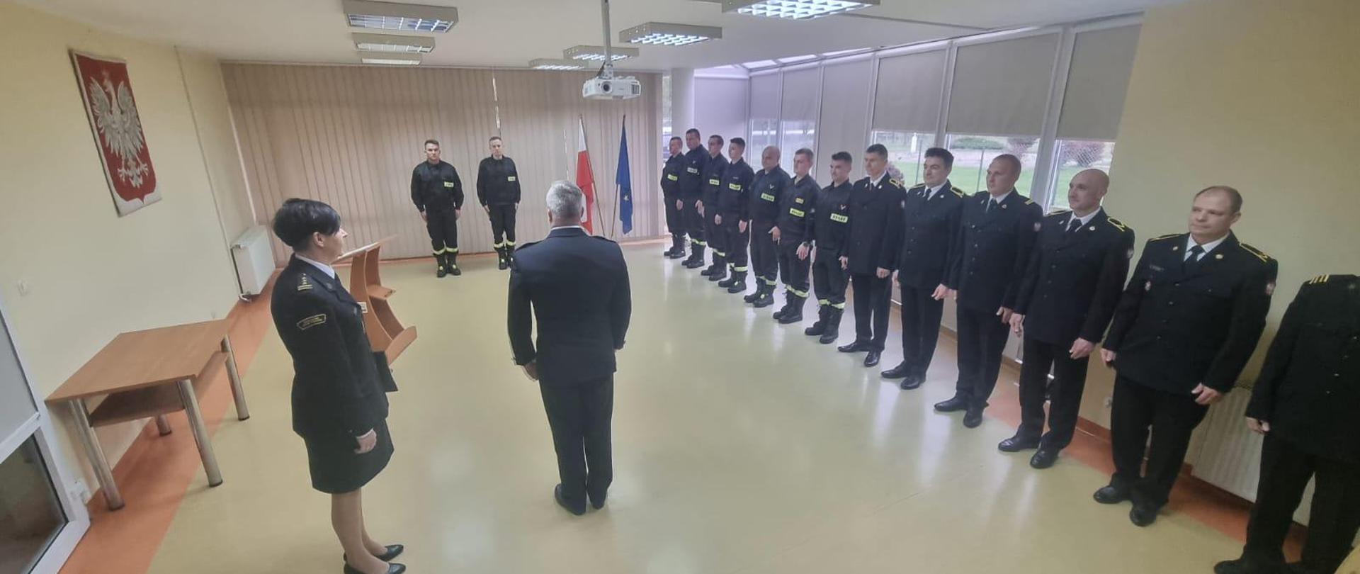 Sala konferencyjna, zgromadzeniu funkcjonariusze podczas uroczystości złożenia ślubowania przez dwóch strażaków. Po lewej stronie widoczne godło po prawej ustawiony szereg strażaków. Na planie głównym Pani Komendant Powiatowa Państwowej Straży Pożarnej w Kamiennej Górze oraz Dowódca jednostki Ratowniczo Gaśniczej.