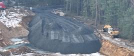 Widoczny czarny materiał przypominający popiół deponowany w wyrobisku górniczym, u góry widać tył odjeżdżającej ciężarówki z prawej strony stoi koparka.