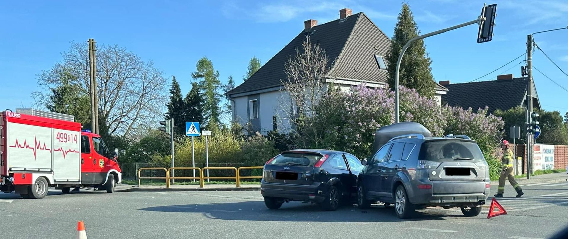 Na zdjęciu znajdują się dwa samochody które się zderzyły na skrzyżowaniu, samochód strażacki oraz strażak. W tle widać budynek