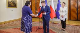 Minister Jan Grabiec wręcza akt powołania do Rady KSAP prof. Jolancie Itrich-Drabarek, obok stoi szefowa służby cywilnej Anita Noskowska-Piątkowska; w tle flagi Polski i Unii Europejskiej