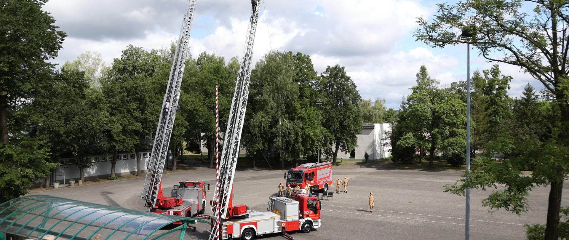 Widok z okna na plac wewnętrzny Szkoły na którym rozstawiono dwa samochody bojowe typu drabina z drabiną wyciągniętą na wysokość 21 metrów po której wchodzą kandydaci zdający egzamin. W tle dodatkowy samochód gaśniczy służący do asekuracji lin, króre zabezpieczają osoby wspinające się po drabinie