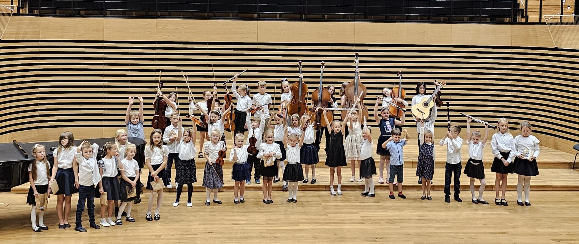 Około czterdzieścioro elegancko ubranych dzieci stoi w trzech rzędach na estradzie sali koncertowej, niektórzy trzymają upominki, niektórzy swoje instrumenty podniesione do góry.