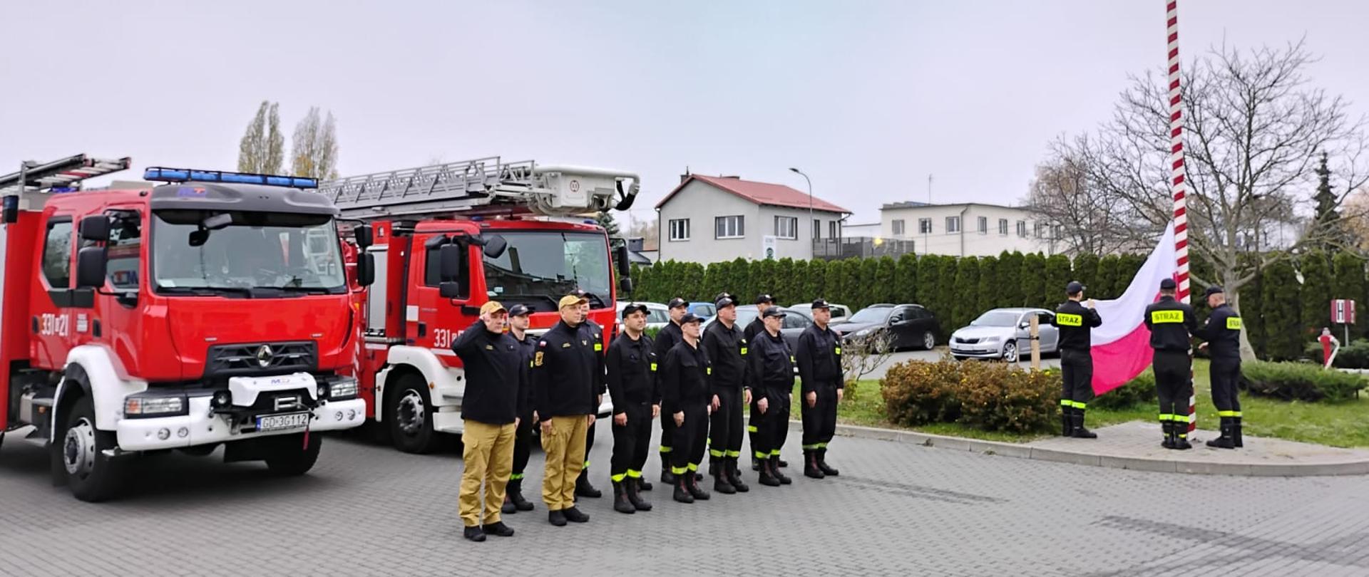 Zdjęcie przedstawia strażaków na tle wozów strażackich podczas uroczystej zmiany służby i podniesienia flagi państwowej na maszt.