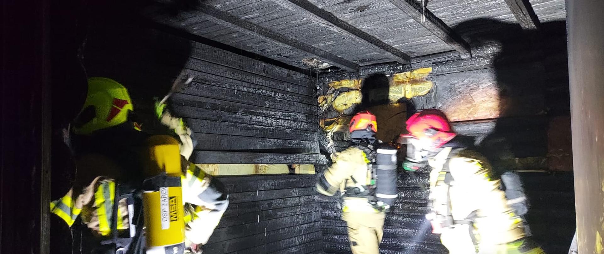 Spalone pomieszczenie, na ścianach i suficie widać zwęglone deski. Trzech strażaków w aparatach oddechowych szuka zarzewi ognia.