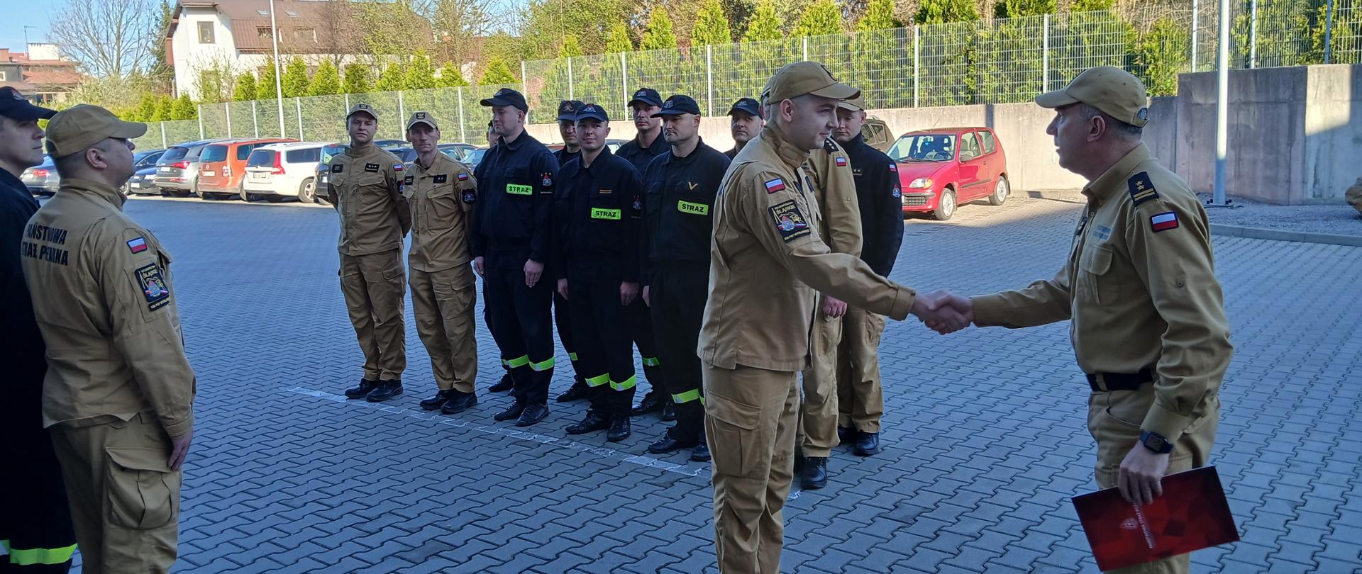 Strażacy stoją na placu wewnętrznym KM PSP Mysłowice w 2 rzędach naprzeciwko siebie. W tle komendant przy zebranych strażakach gratuluje wyróżnionemu strażakowi stojącemu przed nim za wzorowe zachowanie i uratowanie życia ludzkiego w czasie wolnym od służby. 