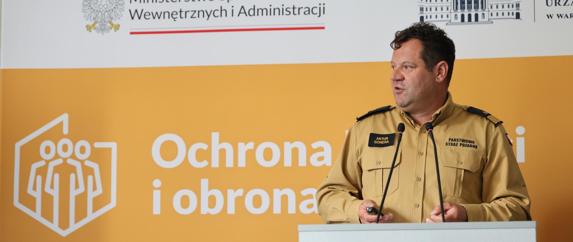 Na zdjęciu Mazowiecki Komendant Wojewódzki PSP nadbryg. Artur Gonera przemawia zza mównicy. Ubrany jest w piaskowy mundur służbowy Państwowej Straży Pożarnej z dystynkcjami i emblematami formacji. W tle znajduje się pomarańczowa ścianka z napisem „Ochrona i obrona” oraz logotypami Ministerstwa Spraw Wewnętrznych i Administracji i Mazowieckiego Urzędu Wojewódzkiego. Zdjęcie oddaje atmosferę profesjonalnego i merytorycznego wystąpienia.