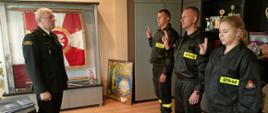Troje strażaków składa ślubowanie w przed Komendantem Powiatowym PSP w Rypinie starszym brygadierem Grzegorzem Łydkowskim. Znajdują się w pomieszczeniu. Na podłodze brązowe panele. W tle po prawo siwe meble, w tle na wprost gablota ze sztandarem.