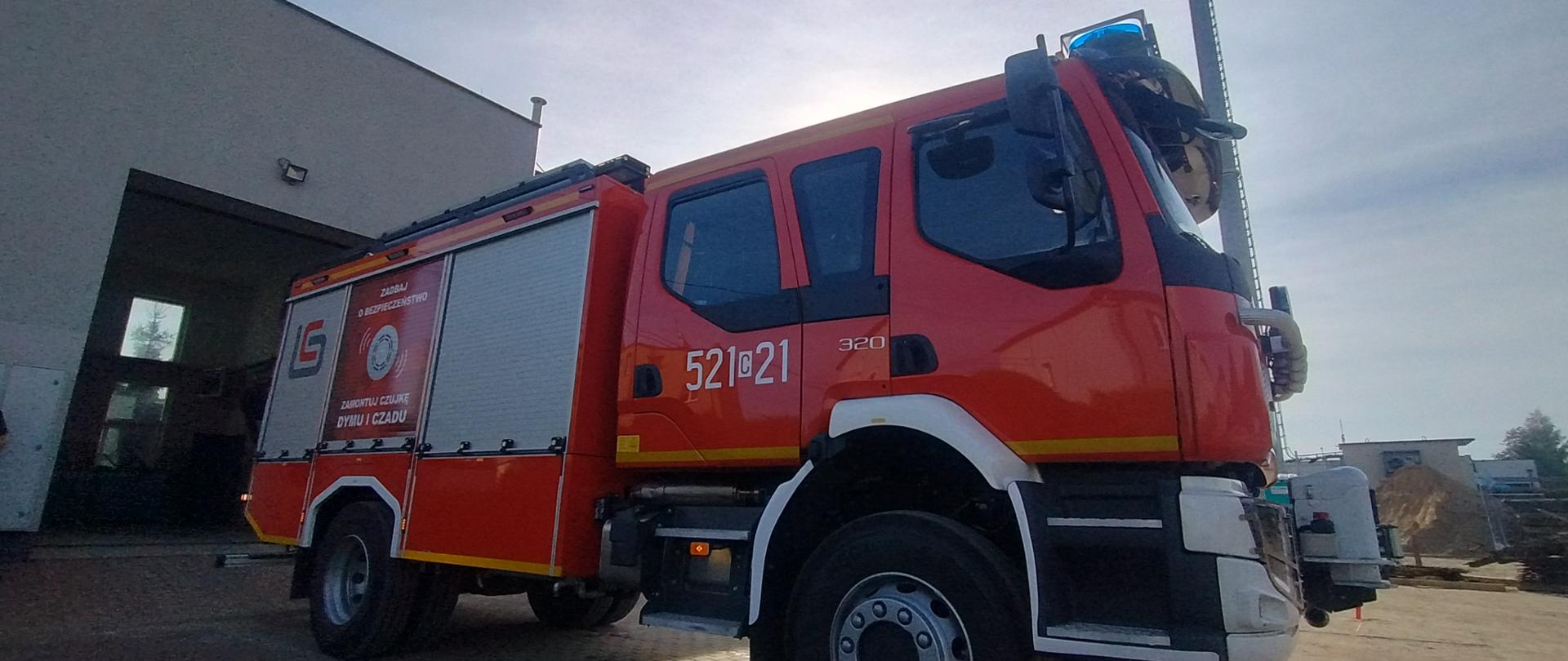 Samochód strażacki ujęty z dolnej perspektywy od strony kabiny pasażerskiej. Na drzwiach widnieje numer operacyjny 521[C]21 oraz oznaczenie modelu 320. Wóz stoi przed garażem remizy strażackiej przy słonecznej pogodzie.
