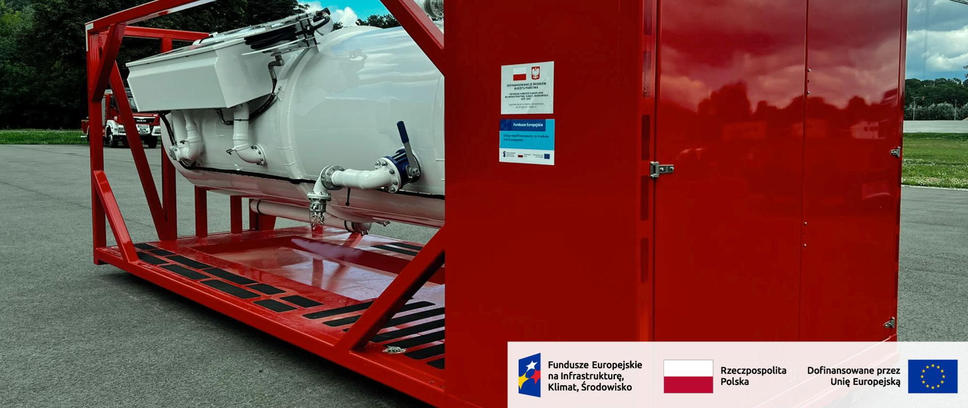 Widok z boku na białą cysternę szkoleniową zamontowaną w czerwonej konstrukcji trenażera. Po prawej stronie znajduje się zamknięty przedział techniczny, a na obudowie tablice informujące o dofinansowaniu. W prawym dolnym rogu oznaczenia Funduszy Europejskich na Infrastrukturę Klimat Środowisko, Rzeczypospolitej Polskiej i Unii Europejskiej.